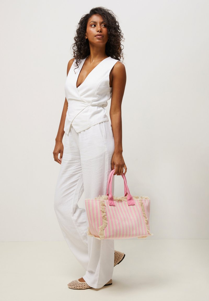 Woman in sleeveless white top and pants holding a pink and beige striped tote bag with fringe detailing, standing against a plain light background.