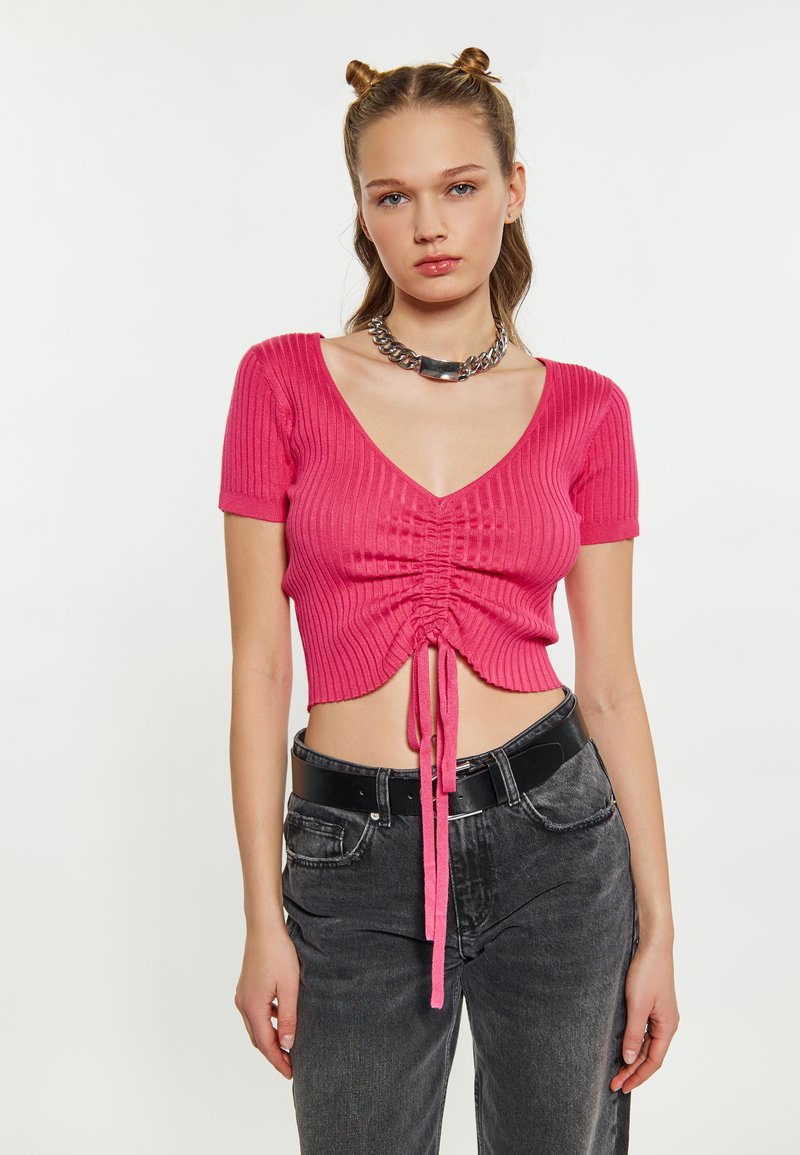 Růžový žebrovaný crop top s krátkými rukávy, s šňůrkou ve středu. Nosený s černými džíny vysokého pasu a černým opaskem.