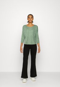 Sweater verde com textura, decote em forma de barco e mangas três quartos, apresentando um padrão de manchas brancas. Usado com calças pretas largas.