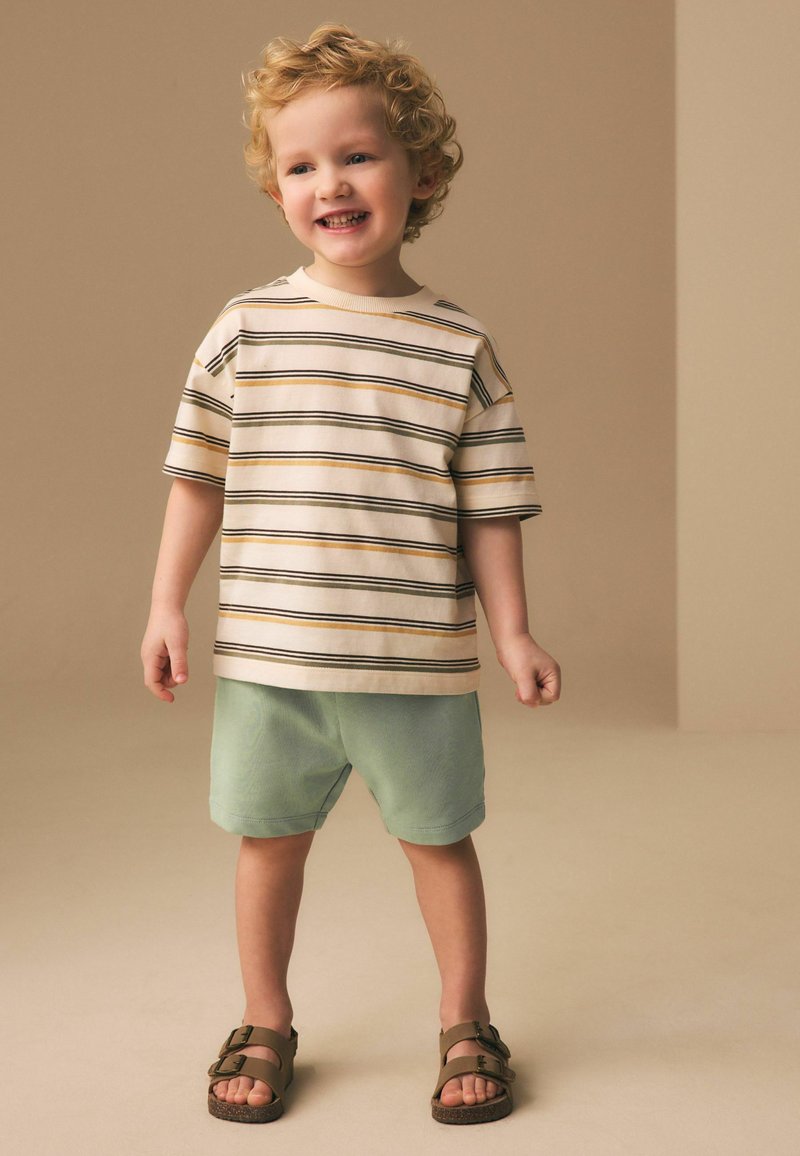 Jeune garçon souriant aux cheveux bouclés portant un t-shirt à manches courtes rayé, un short vert et des sandales marron, debout sur un sol beige.