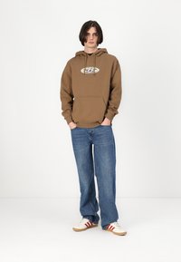 Sweat-shirt à capuche marron avec une poche avant et un logo, associé à un jean bleu ample et des chaussures blanches avec des rayures rouges.
