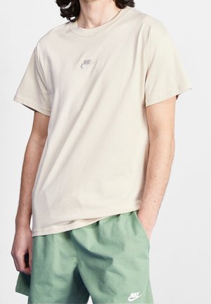 Personne portant un t-shirt Nike beige clair et un short Nike vert, debout avec une main dans la poche devant un fond uni.