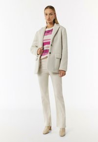 Weißer strukturierter Blazer mit zwei Knöpfen, getragen über einem rosa-weißen gestreiften Pullover. Cremefarbene Schlaghose und beige Blockabsatzschuhe.