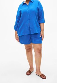 Loose-fitting blue shirt with rolled sleeves, paired with matching blue shorts. Soft textured fabric, worn with brown flat sandals.