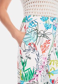 Die Blumenmusterhose hat einen weißen Hintergrund mit bunten, handgezeichneten Pflanzendesigns in Blau, Grün, Rot und Pink. Eine Seiten Tasche ist sichtbar.