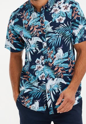 Man draagt een kortermig navyblauw overhemd met knopen met een tropisch patroon van witte vogels, bloemen en blauwe en oranje bladeren.