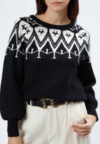 Schwarzer Strickpullover mit einem weißen geometrischen Muster am Schulterbereich, mit überschnittenen Schultern und einer strukturierten Oberfläche. Kombiniert mit cremfarbenen Hosen und einem dekorativen Gürtel.