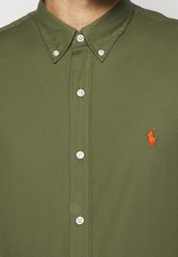 Koszula khaki na guziki wykonana z bawełnianej tkaniny, z ostrokątnym kołnierzem, białymi guzikami oraz pomarańczowym, wyszywanym logo na piersi.