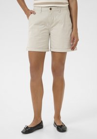 Kvinna i beige shorts med uppvikt fåll, randig topp och svarta balettskor som står med händerna i fickorna mot en vit bakgrund.