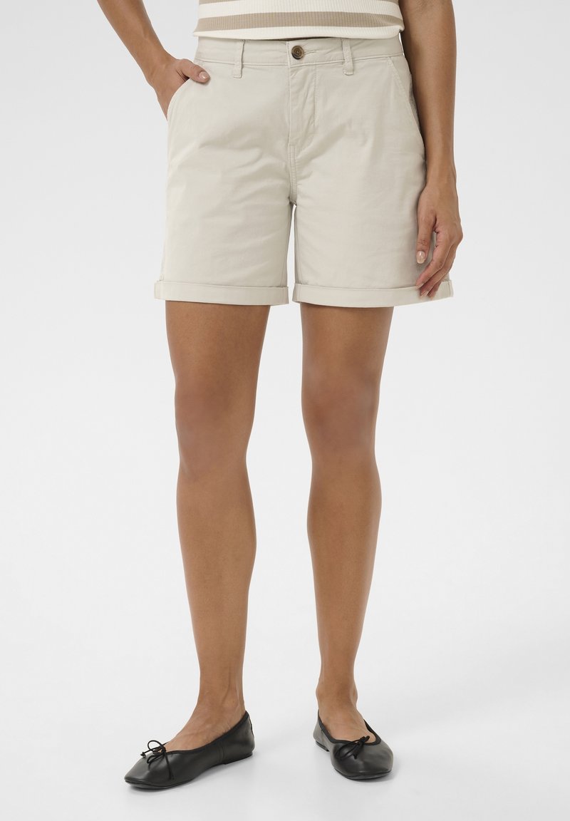 Kvinna i beige shorts med uppvikt fåll, randig topp och svarta balettskor som står med händerna i fickorna mot en vit bakgrund.