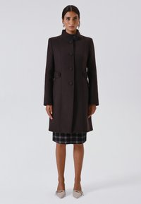 Vrouw in een donkerbruin knopenjasje met staande kraag, geruite rok, beige puntige hakken en grote ringoorbellen, staande tegen een effen achtergrond.