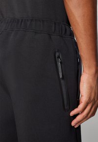 Gros plan d'une personne portant un pantalon noir avec une taille élastique et une poche zippée verticale sur le côté près de la main.