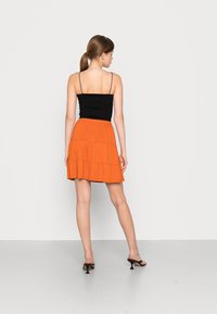 Camisola preta ajustada e uma saia mini em camadas laranja fluida, feita de tecido suave. O conjunto é combinado com sandálias de salto pretas.