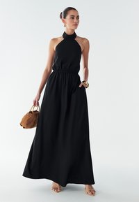 Črna maxi obleka s halter ovratnikom in nabranim pasom, z bočnima žepoma. Kombinirana s rjavo semiš torbo in bež sandali.