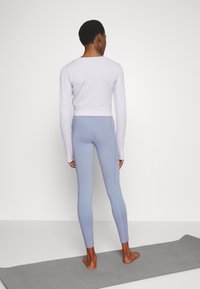 Top a maniche lunghe corto di colore viola chiaro abbinato a leggings blu chiaro. Il tessuto ha una texture liscia e presenta dettagli di cucitura a contrasto.