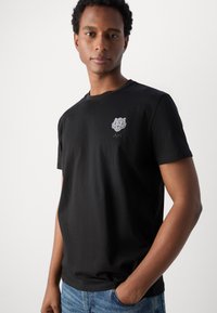 T-shirt en coton noir avec un graphisme de tigre sur la poitrine gauche et les initiales "AM", avec une coupe décontractée et des manches courtes.