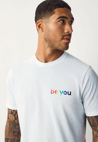 Hvid bomulds t-shirt med multicoloreret "være dig" tekst på venstre bryst. Korte ærmer og en simpel, klassisk rund halsudskæring.