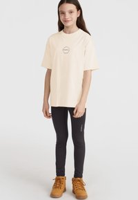 T-shirt en coton beige clair avec un logo circulaire, associé à des leggings noirs et des bottes jaunes avec des lacets marron et des semelles texturées.
