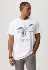 Biała bawełniana koszulka z grafiką postaci z skrzydłami oraz napisem "City of Angels" w niebieskim kolorze; noszona z jasnoszarymi spodniami.