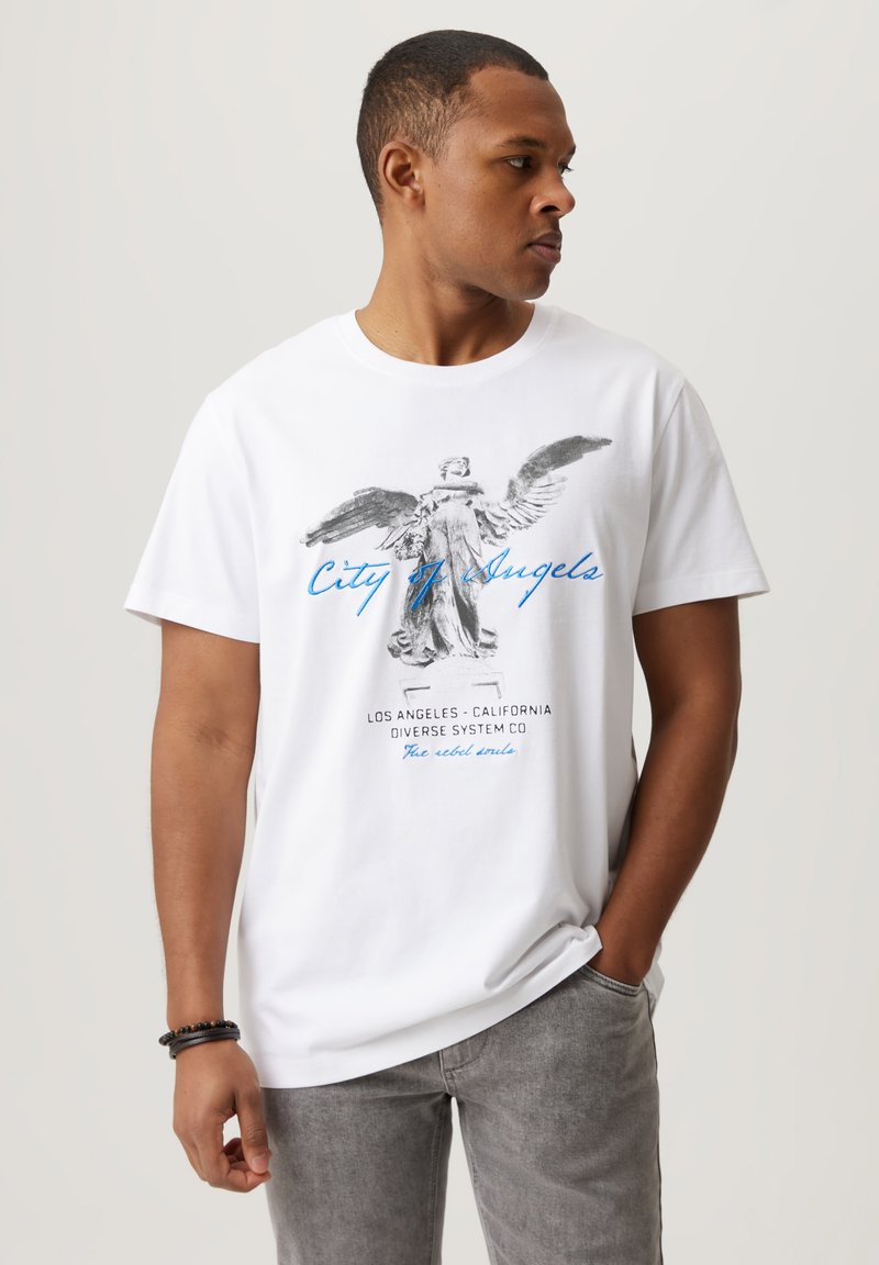 Biała bawełniana koszulka z grafiką postaci z skrzydłami oraz napisem "City of Angels" w niebieskim kolorze; noszona z jasnoszarymi spodniami.