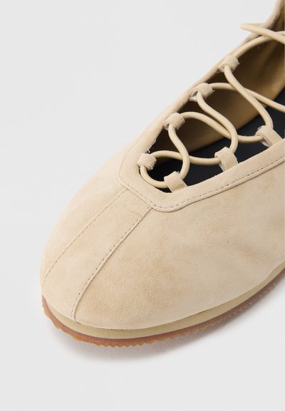 Μπεζ παπούτσι από σουέντ με στρογγυλεμένη μύτη, με ελαστικά κορδόνια που διασταυρώνονται και υφή στη σόλα από καουτσούκ. Ομαλή επάνω επιφάνεια με ραμμένες λεπτομέρειες.