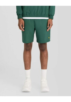 Mand iført mørkegrønne shorts med et lille gult logo på venstre ben, hvide sokker og hvide sneakers, står foran en ensfarvet baggrund.