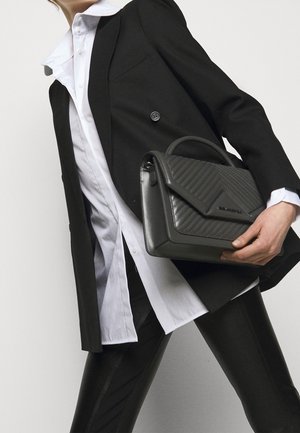 Personne portant un blazer noir et une chemise blanche tient un sac à main en cuir matelassé noir avec une poignée supérieure sur un fond uni.