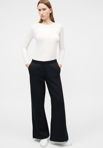 Γυναίκα που στέκεται με τα χέρια στις τσέπες, φορώντας λευκό μπλουζάκι με μακριά μανίκια, μαύρο παντελόνι με φαρδιά μπατζάκια και λευκά αθλητικά παπούτσια σε απλό φόντο.