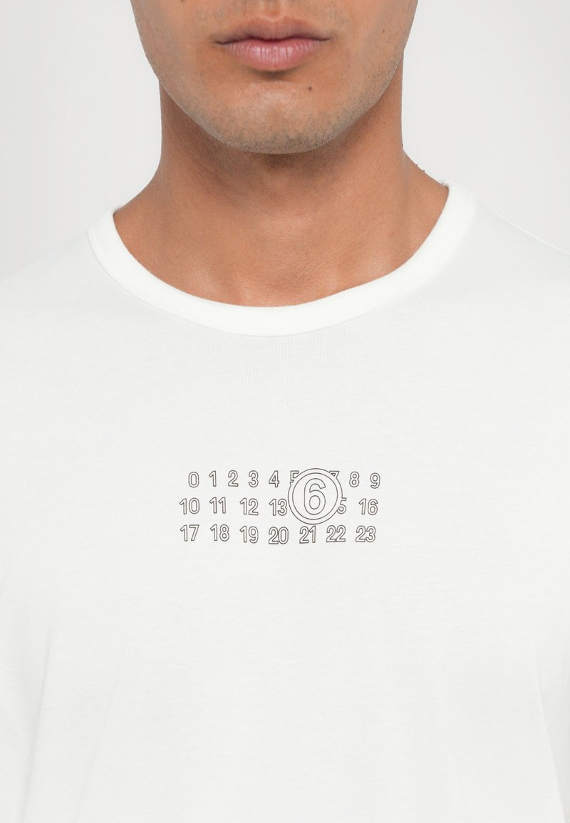 T-shirt bianco in cotone con un design stampato con numeri, inclusa una '6' evidenziata in un cerchio al centro. Maniche corte e scollatura rotonda.