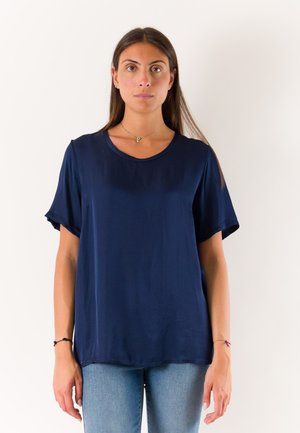 Donna in piedi, rivolta in avanti, che indossa una camicetta semplice blu scuro a maniche corte e jeans azzurri su sfondo bianco.