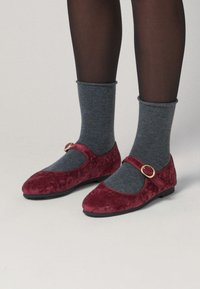 Zapatos Mary Jane de terciopelo burdeos con puntera redondeada y una correa con un hebilla circular dorada, combinados con calcetines tobilleros de algodón gris acanalados.