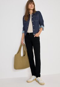 Giacca di jeans con due tasche frontali, polsini azzurri; maglione chiaro; pantaloni neri; borsa verde oliva; scarpe da ginnastica bianche con dettagli blu.
