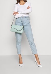 Een mintgroene handtas met krokodillenstructuur, een gebogen klep en zilverkleurige details, gecombineerd met lichtblauwe high-waisted jeans en witte hakken.