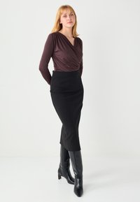 Haut à manches longues couleur bordeaux profond avec un design croisé, associé à une jupe noire cintrée et des bottes noires montant jusqu'aux genoux. Tissu lisse, silhouette élégante.