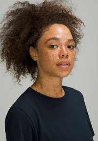 Een persoon met krullend, bruin haar en sproeten draagt een donkerblauw t-shirt. De achtergrond is eenvoudig, waardoor de kenmerken van het onderwerp extra nadruk krijgen.