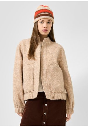 Jeune femme portant une veste beige texturée, un bonnet rayé en tricot et une jupe marron à boutons sur un fond clair uni.