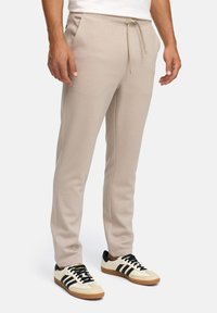 Beige drawstring sweatpants worn with cream sneakers featuring black stripes and gum soles, shown on a standing male model.