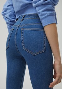 Hubené modré džíny z denimu, s vysokým pasem a dvěma zadními kapsami s kontrastním šitím. Textura vypadá hladce.