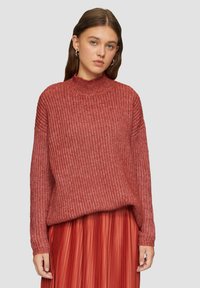 Czerwony sweter dzianinowy z wysokim kołnierzem i luźnym krojem, z teksturowanym wzorem i ściągaczami, w połączeniu z plisowaną czerwoną spódnicą.
