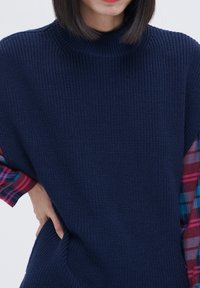 Tummansininen, ribattu neulepusero, jossa on korkea kaulus ja ruutukangasviimeistely hihoissa. Sisältää sivutaskun.