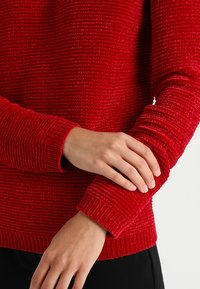Červený svetr s texturou a žebrovaným vzorem, s dlouhými rukávy a kulatým výstřihem. Materiál je jemný a plyšový.