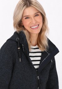 Tamsiai mėlynas užtraukiamas džemperis su tekstūrine medžiaga, su aukštu apykakle, reguliuojama virvele ir sidabro spalvos detalėmis, dėvimas ant dryžuotos marškinėlių.