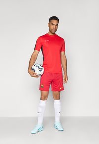 Punane Nike jalgpallipaita mustade aktsentidega, punased lühikesed püksid FC Barcelona logoga, valged sokid ja helesinised tossud, hoides jalgpalli.