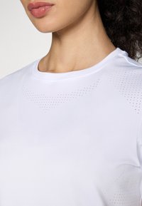 Valge sportlik särk, mis on valmistatud kerge kangaga, millel on ümmargune kaelus ja väikesed aukudega perforatsioonid hingavuse tagamiseks õlgadel.