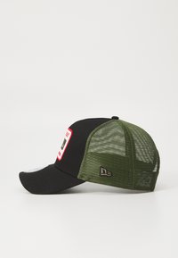 Gorra negra con un panel frontal de malla en verde oliva. Presenta un parche rectangular con texto en rojo y negro, y un logotipo de la marca en el lateral.