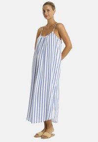 Una donna in profilo indossa un vestito estivo a righe blu e bianche con spalline sottili, abbinato a sandali beige. La luce naturale mette in risalto i suoi lineamenti.