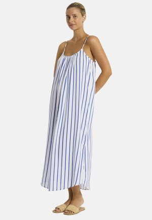 Eine Frau steht im Profil und trägt ein gestreiftes blau-weißes Sommerkleid mit dünnen Trägern, kombiniert mit beigen Slipper-Sandalen. Das natürliche Licht betont ihre Gesichtszüge.
