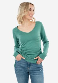 Top a maniche lunghe, con scollatura a V, realizzato in tessuto teal con dettagli arricciati sui lati e maniche aderenti. Abbinato a jeans blu. Texture liscia.