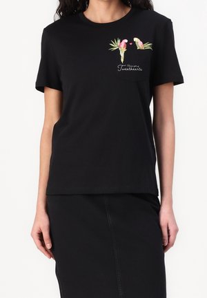 Femme portant un t-shirt noir avec des perroquets roses brodés, des feuilles vertes, un petit cœur et un texte indiquant "Petsafe club by Tweethearts."