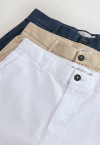 Trois pantalons chino pliés en blanc, beige et bleu marine, chacun avec des passants de ceinture et un bouton noir à la taille.